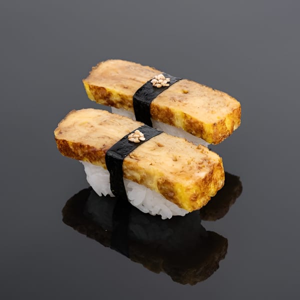 Two pieces of tamago nigiri sushi with white rice, egg omelet, nori, and sesame seeds.