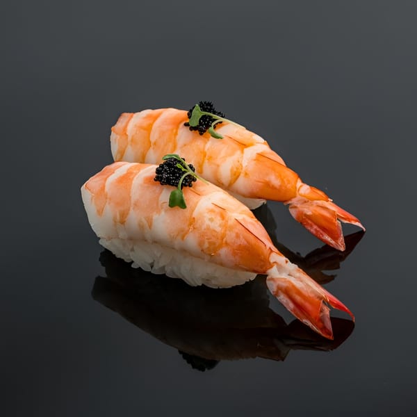 Two shrimp nigiri sushi pieces, topped with black caviar and microgreens, on a dark reflective surface.
