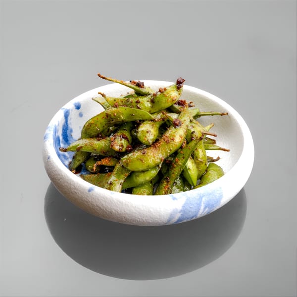 A white bowl with blue accents filled with seasoned edamame pods on a gray surface.